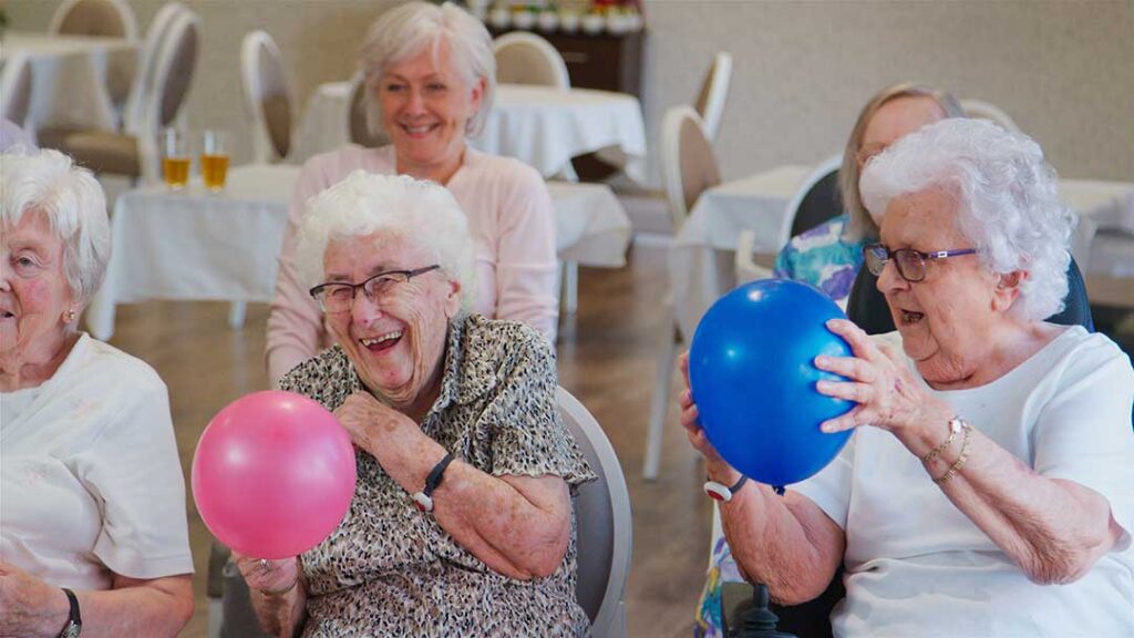 Smiling older adults chatting together in a comfortable, sunlit lounge area with caregiver nearby.