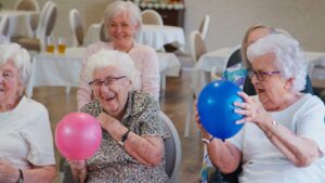 Smiling older adults chatting together in a comfortable, sunlit lounge area with caregiver nearby.